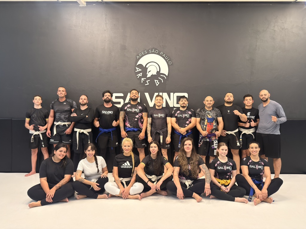 Alunas e equipe da Salvino BJJ reunidas na academia, diante do logo