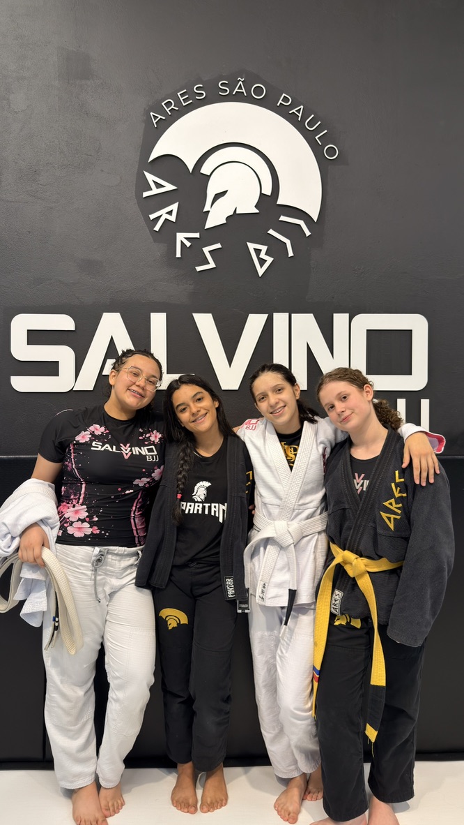 Quatro alunas da Salvino BJJ abraçadas diante do logo da academia